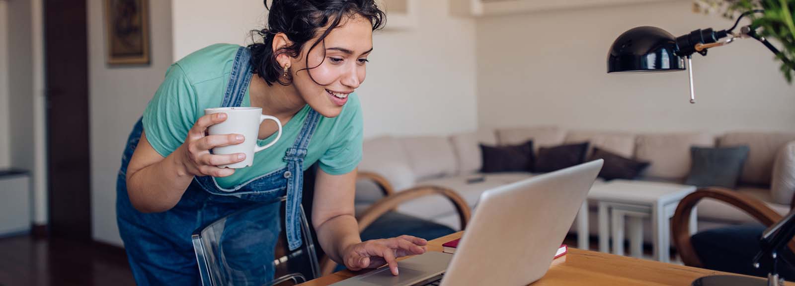 Smiling woman holding coffee mug leans down to view her laptop screen.